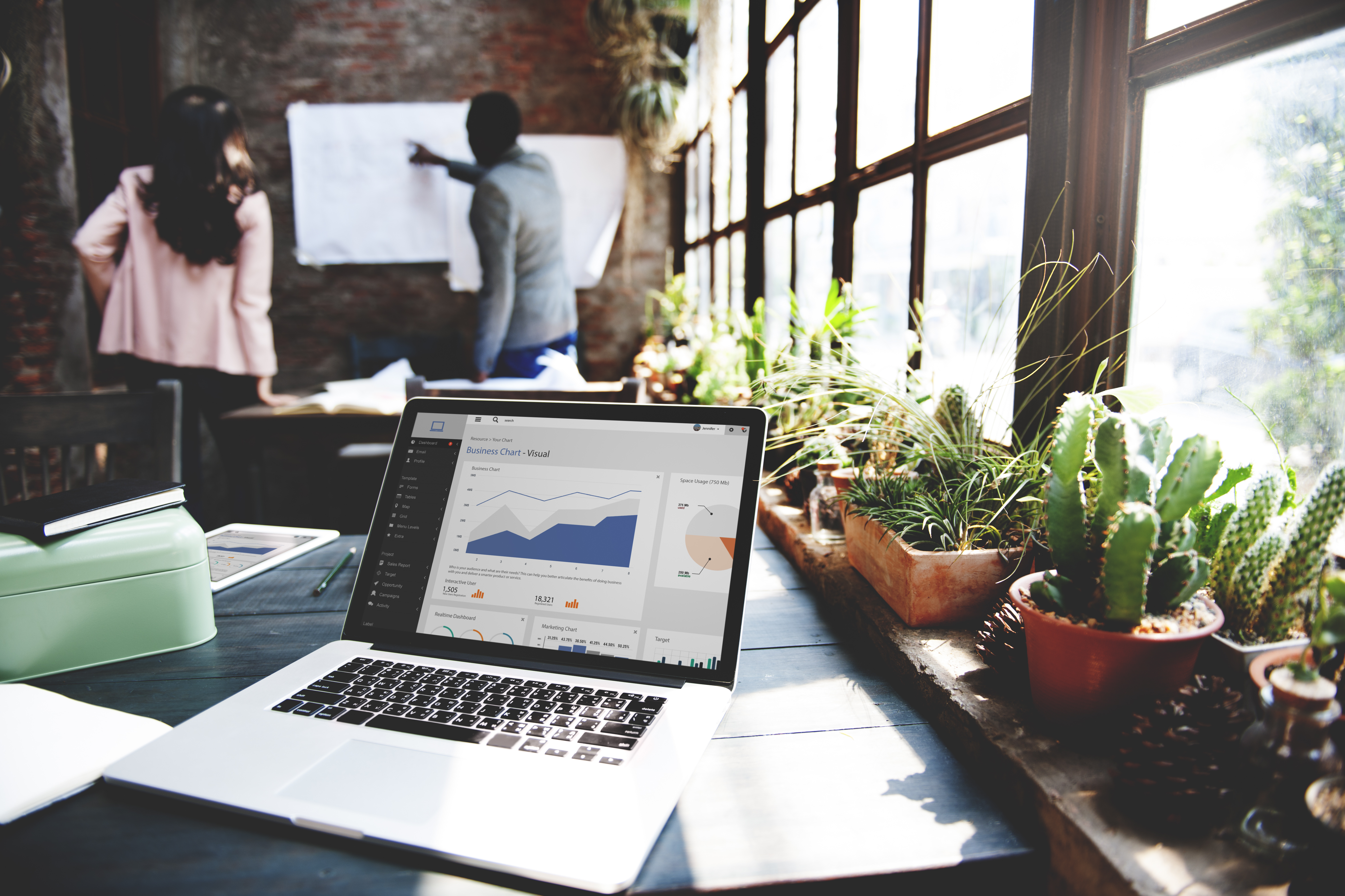 A laptop sits open on a table, the screen displaying a generic business chart template. On the right, a window bathes the various cacti and succulents on the windowsill in bright light as a man and woman pore over a whiteboard in the background.