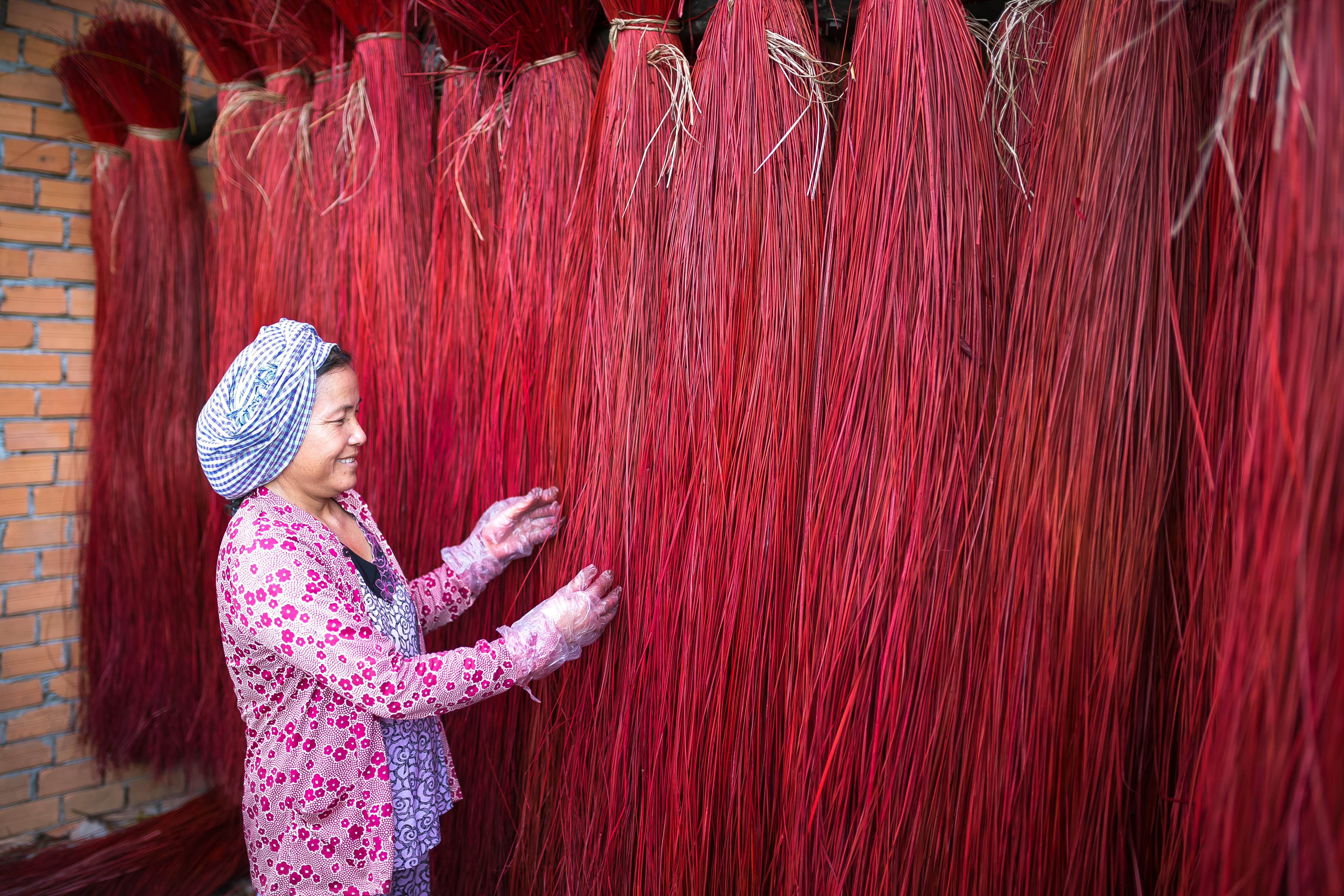 A smiling woman in a pink and blue outfit with a blue and white headscarf and clear plastic gloves handles hanging bundles of red fibres suspended from the ceiling.