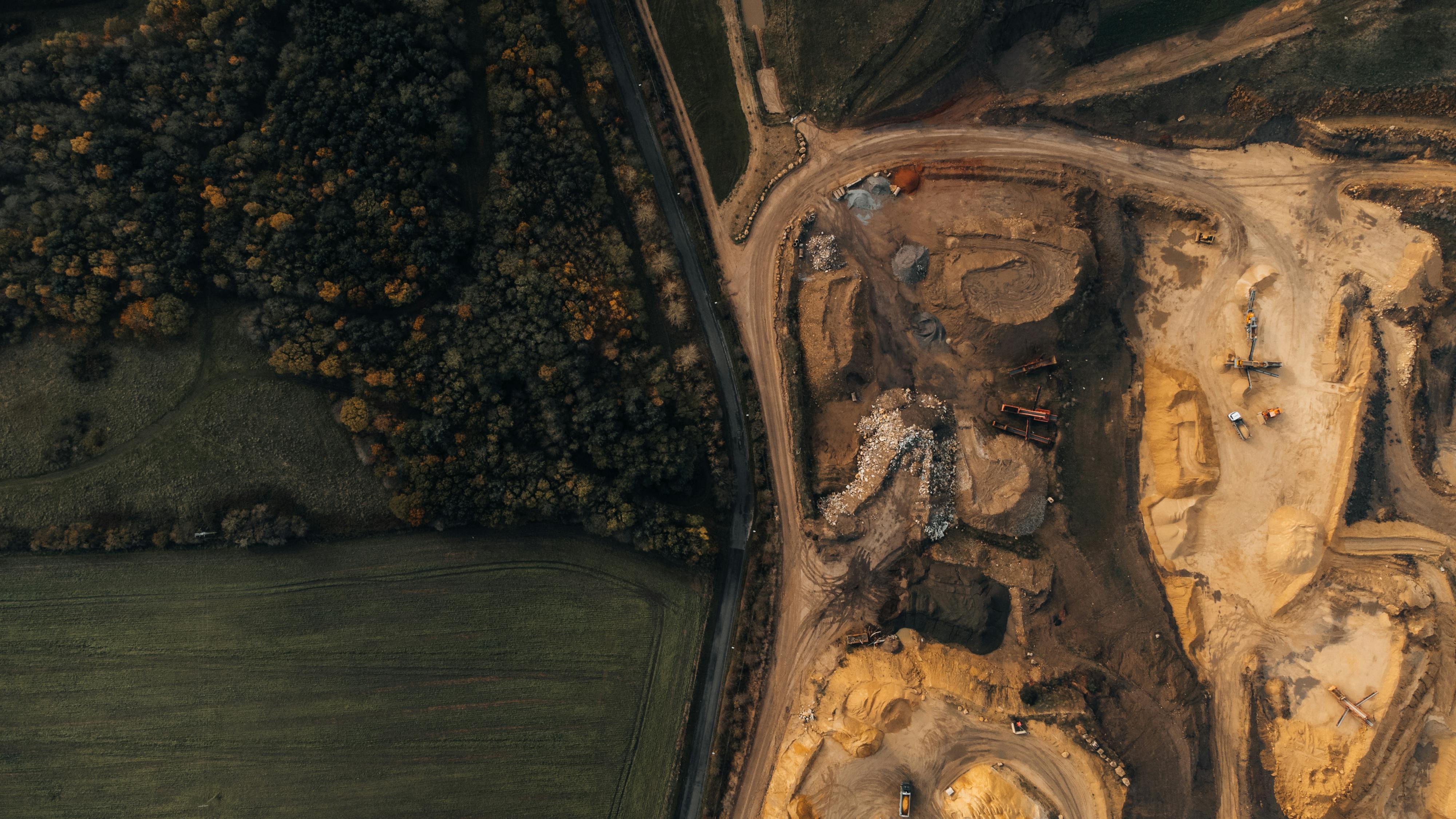 An aerial view of two parallel roads, one tarmac, one yellow dirt, bisecting a green forest and fields on the left side of the image and a bare earth mining site on the right, containing machinery and vehicles.