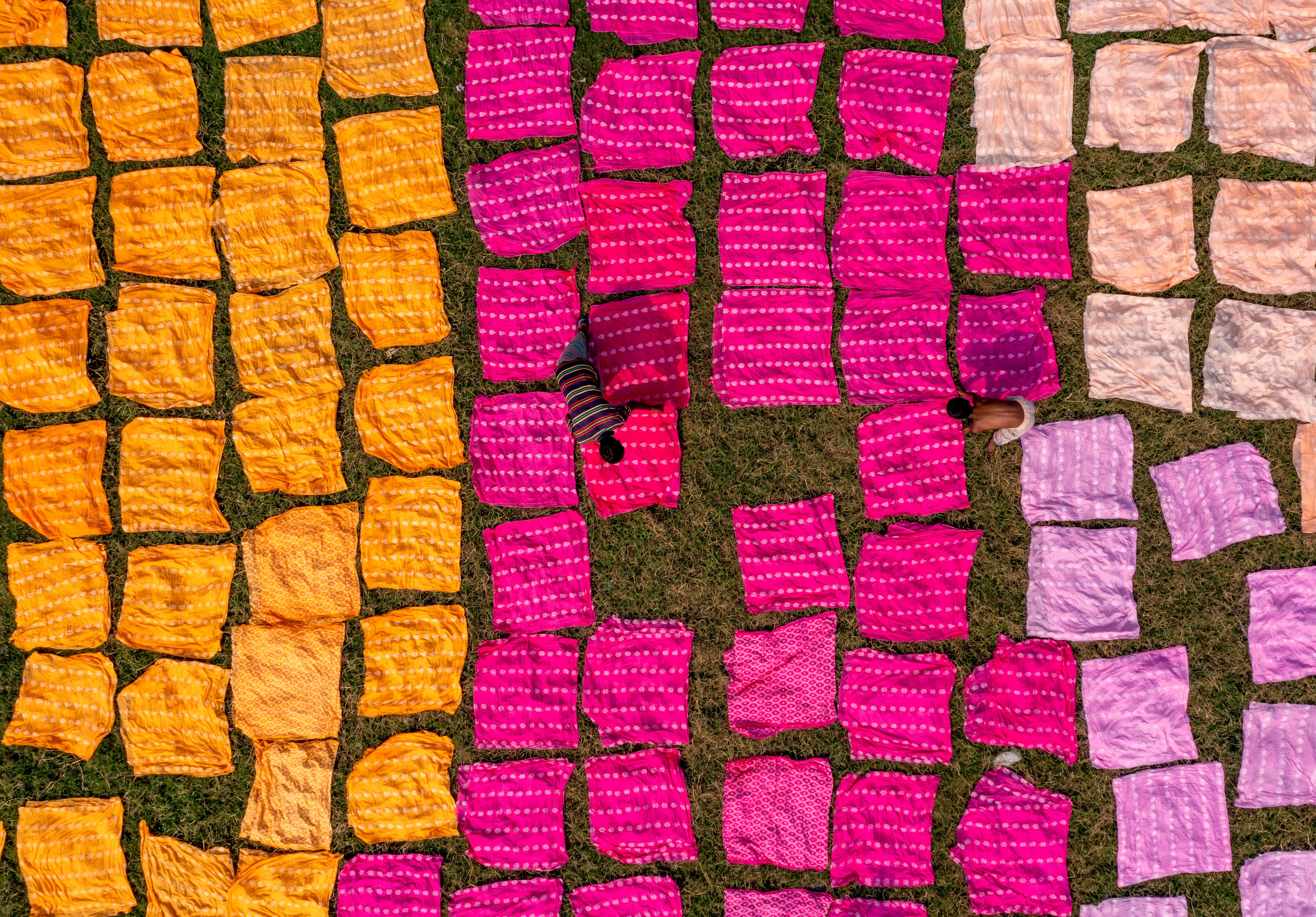 A top-down view of two textile workers amongst sheets of colourful cloth laid flat on a grass field. The sheets are separated by colour, with orange, pink, peach and lavender left-to-right.
