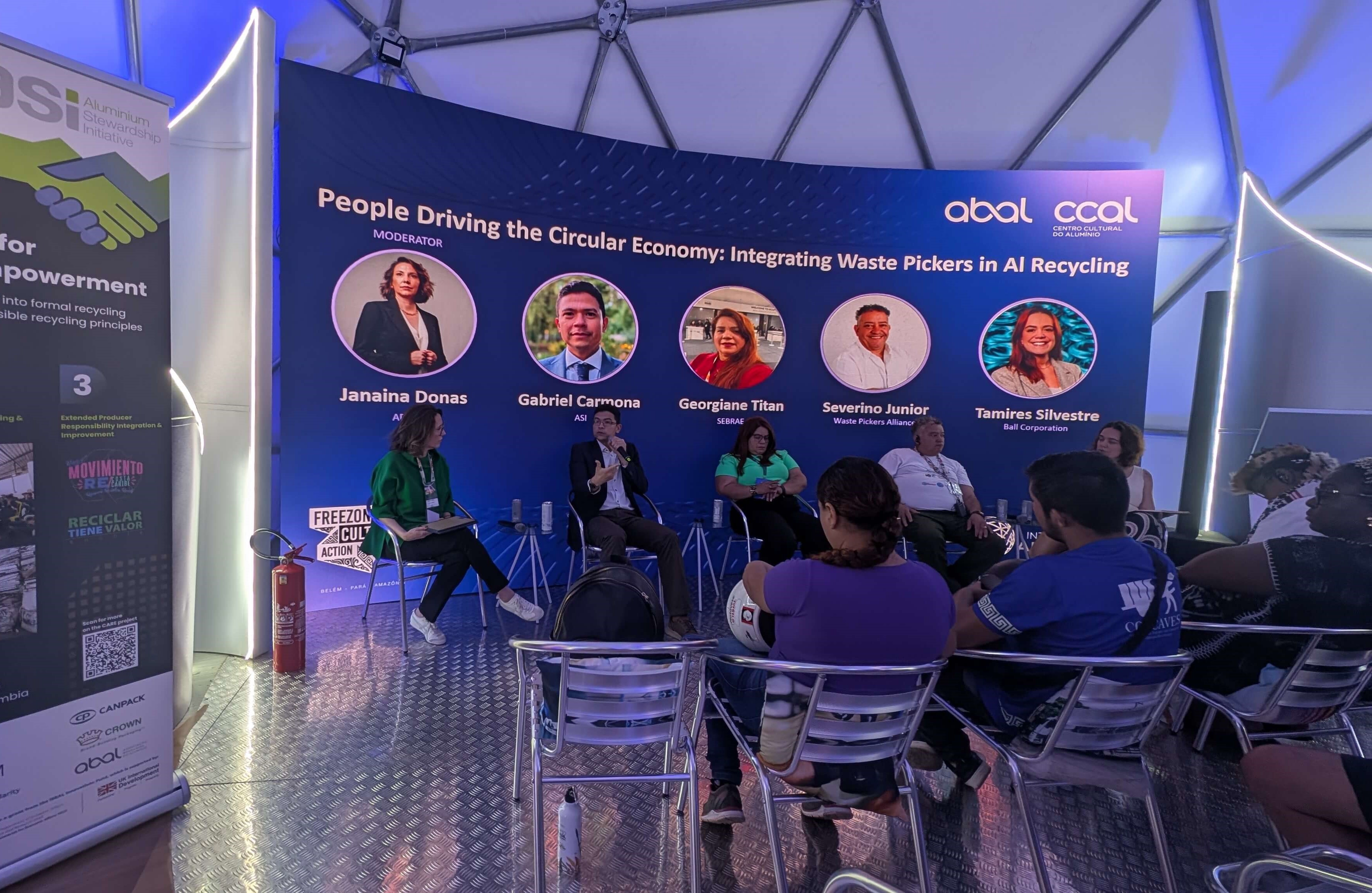 A panel of speakers sit in front of a blue information panel, facing conference attendees. 