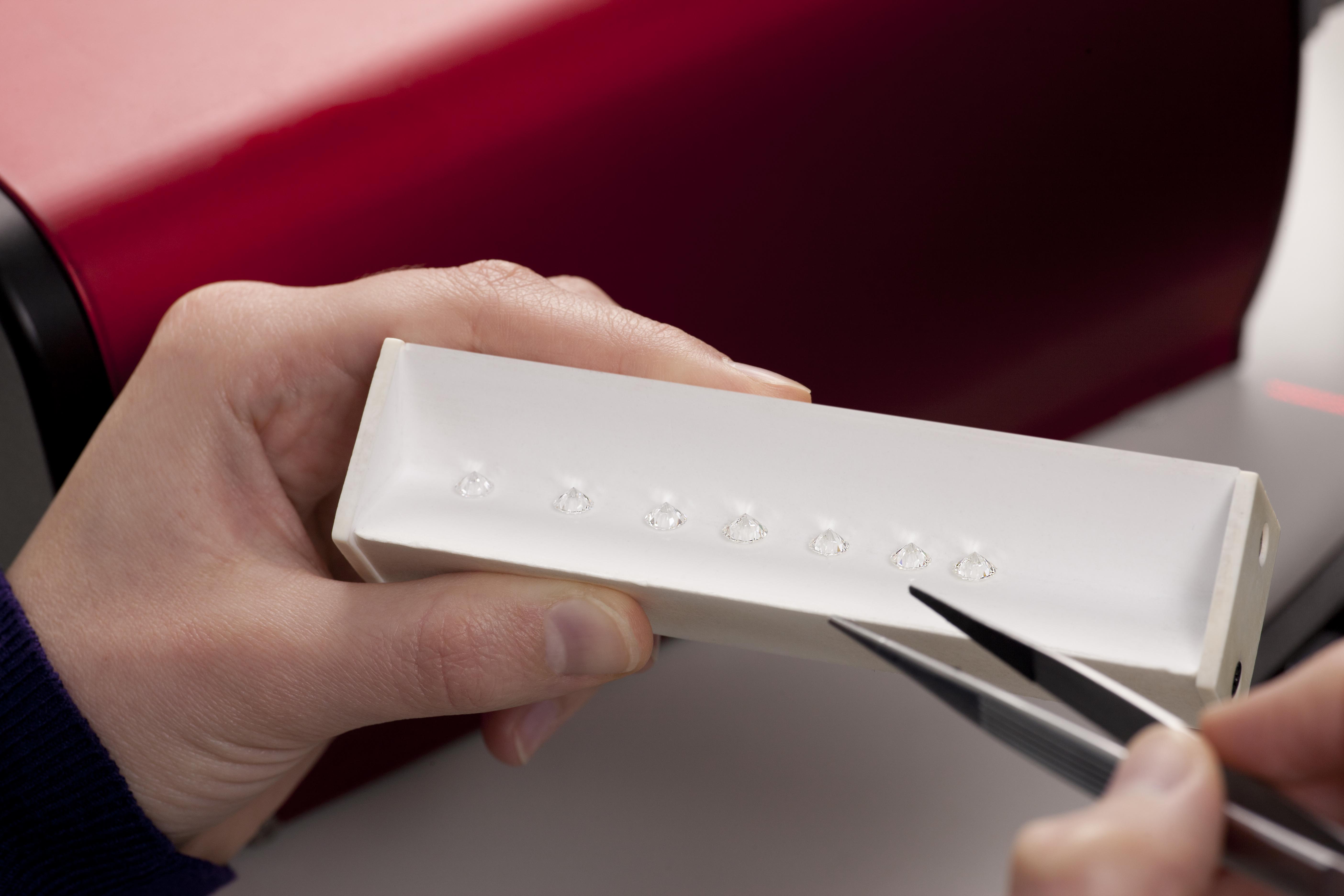 A persons' left hand holds a row of seven clear diamonds in a white jewellers tray, while their right is bringing a set of tweezers closer to the gems.