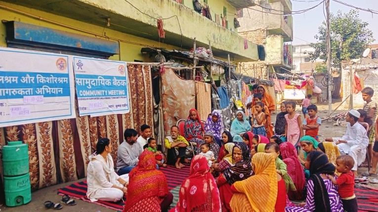 A group of men, women in colourful headscarves, and children are sat and stood in a rough semicircle on a red and blue striped mat in the street, listening to a GoodWeave representative.