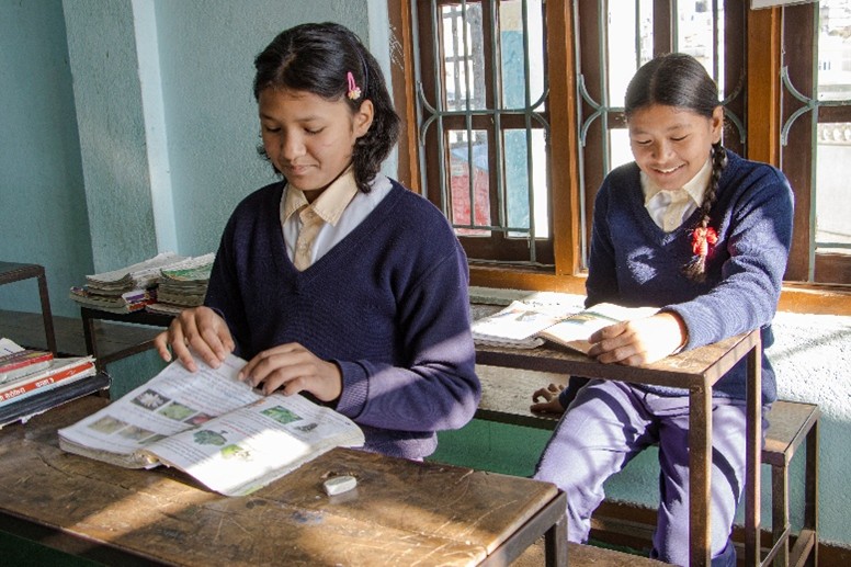 Two smiling Nepalese schoolgirls in navy blue uniforms are reading at their desks in a sunlit classroom.
