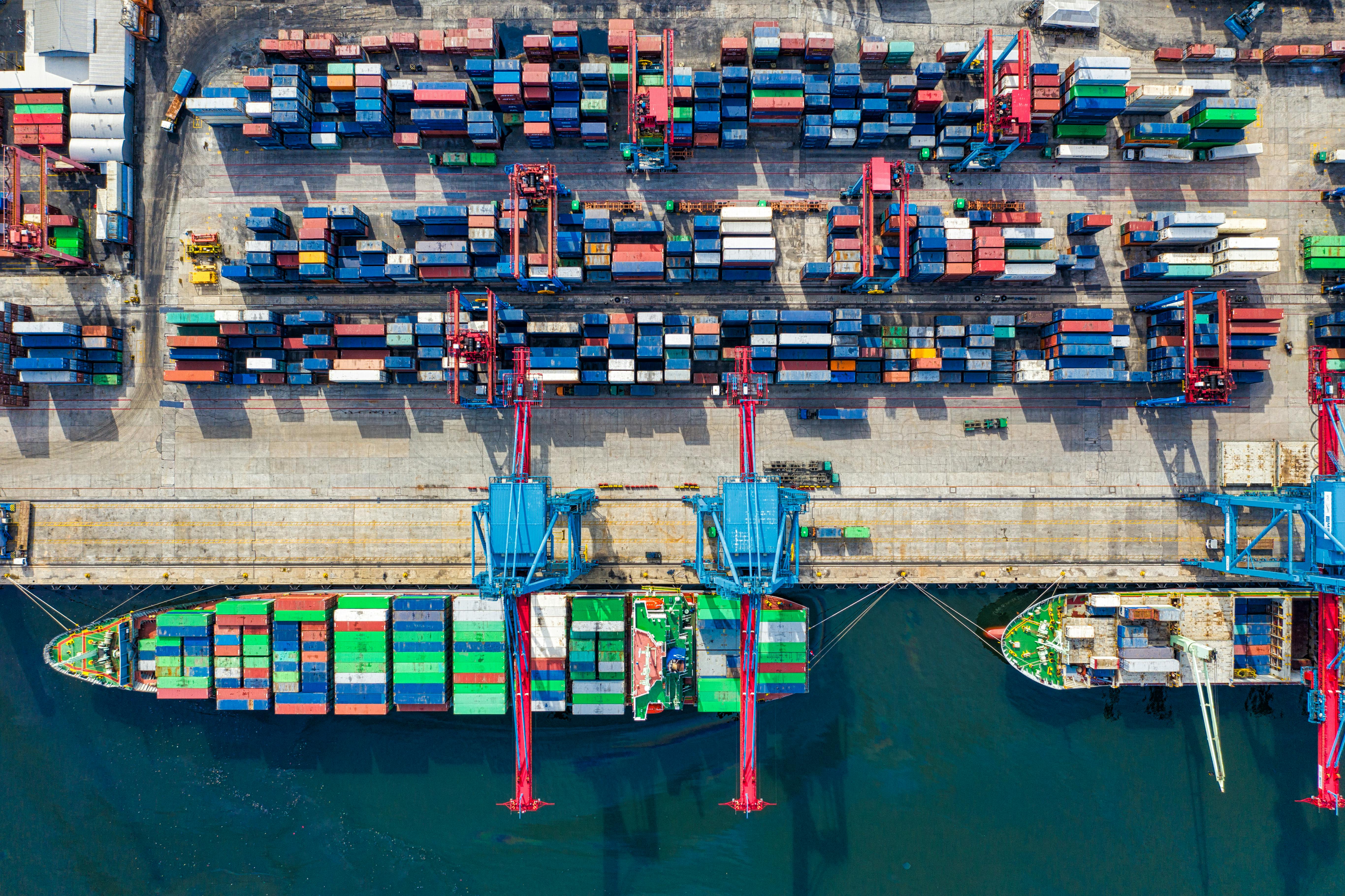 Aerial view of two container ships dockside, showing loading cranes and stacked containers at the waterfront.