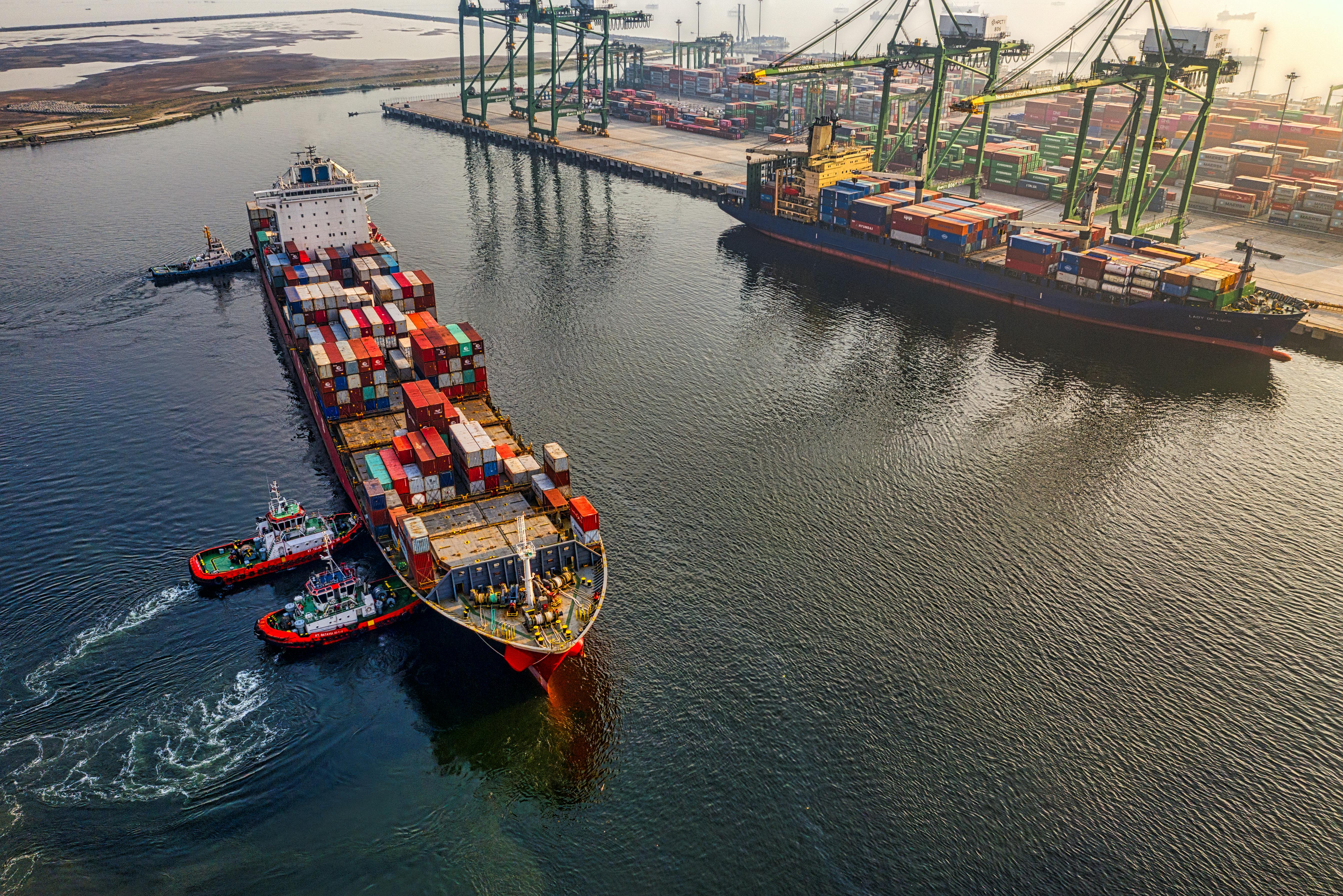 The sun illuminates three tugboats pushing the starboard side of a container ship towards the dock, where another ship, loading cranes and containers await.