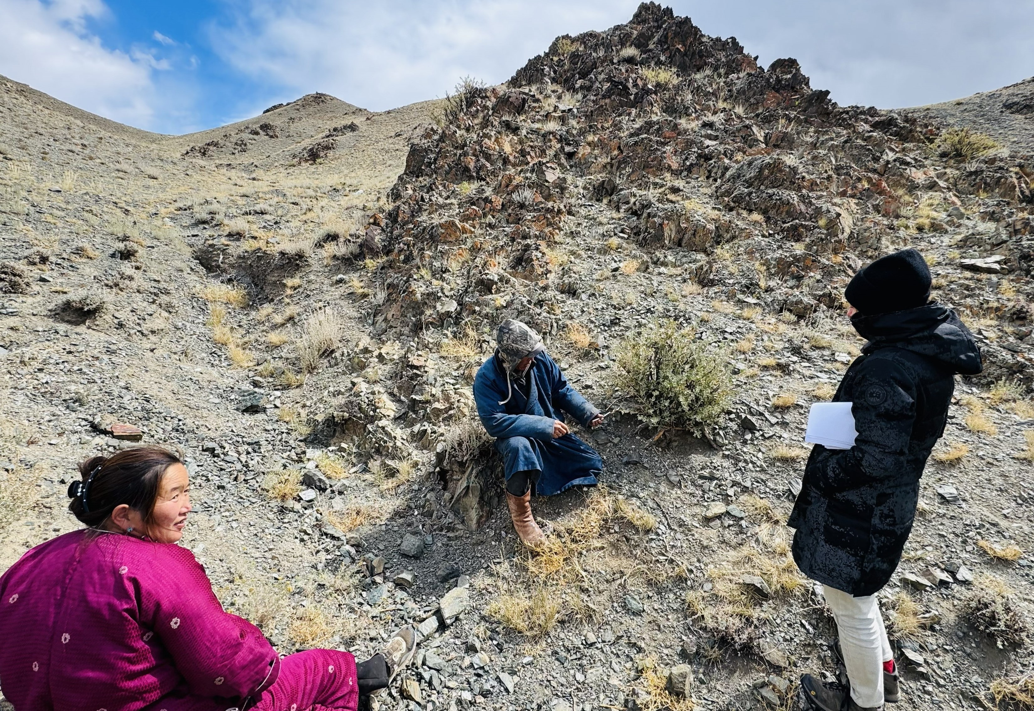 Three people examining soil in an arid, hilly landscape