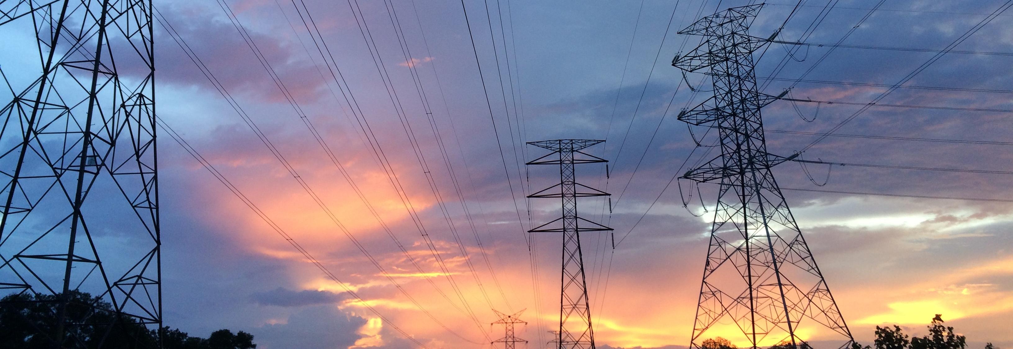 Image of transmission towers against a grey cloudy sky with a yellow sunset at the bottom. Photo by Pok Rie on Pexels