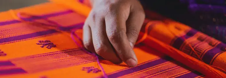 The brown-skinned hand of a textile worker is in focus, resting over loose threads on a bright orange and purple woven cloth