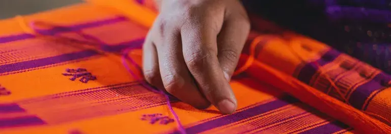 The brown-skinned hand of a textile worker is in focus, resting over loose threads on a bright orange and purple woven cloth.