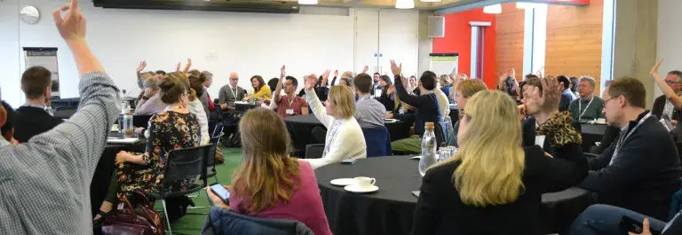 Groups of people sitting around tables listening to a speaker with their hands up