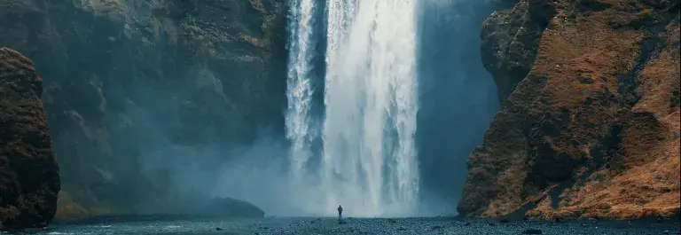 Large waterfall surrounded by rocks with a person in the distance standing near it© Adobe Stock