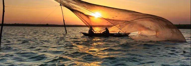 Two people sit silhouetted in a shallow boat on the water at sunset casting an enormous fishing net which floats like a diaphanous membrane above their heads © Pexels