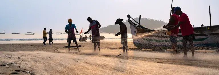 A dawn scene of fishermen working collaboratively, gathering nets on a sunlit coastal shore, with wooden boats docked nearby