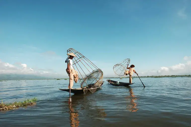 Two people wearing traditional rice hats on small fishing boats with large cages and oars against the blue sky © Unsplash