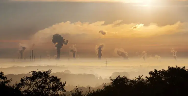 The sun rises over plumes of emissions gas from powerplants dotted along the horizon, which emanate into the sky beyond a smog which covers the trees and houses in the foreground.