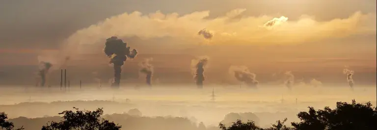 The sun rises over plumes of emissions gas from powerplants dotted along the horizon, which emanate into the sky beyond a smog which covers the trees and houses in the foreground