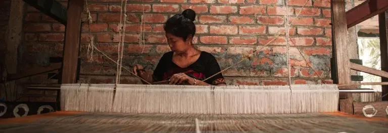 Woman weaving textiles against a red brick wall © Pexels