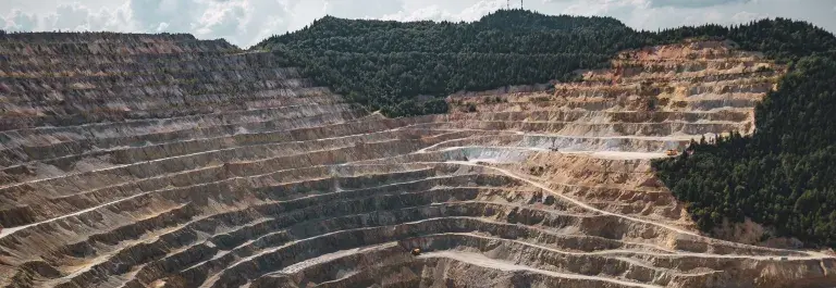A deep, stepped mine site of brown and yellow rock and dust. Roads wind up the sides with mining vehicles moving up and down. Two yellow excavators sit at the bottom next to a pool of blue water. Dense forest mantles the upper edge and right hand shoulder of the site, all under a bright sunny sky with white clouds.