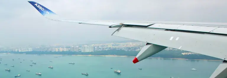 View from an aircraft wing of a number of container ships anchored off a coastline featuring both forest and high rise buildings in a brightly coloured ocean © RSB