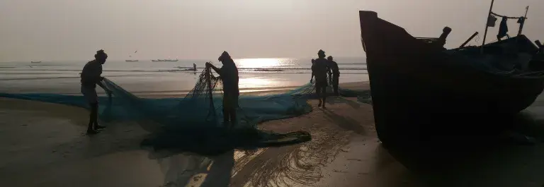 Several fishers pulling a net next to a small boat on a sunny beach