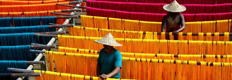 Two people hanging coloured yarns and textiles in the sun