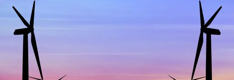 The silhouettes of four wind turbines stand against a blue sky at dusk with an orange sunset on the horizon