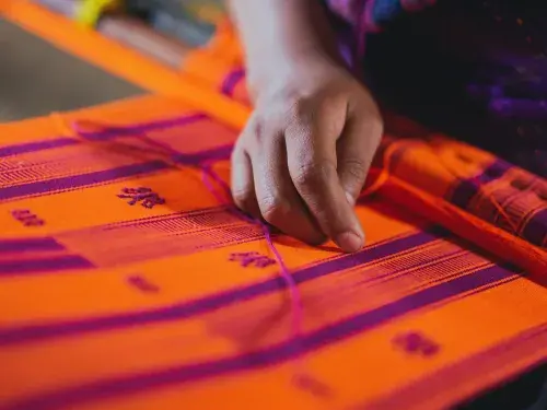 The brown-skinned hand of a textile worker is in focus, resting over loose threads on a bright orange and purple woven cloth