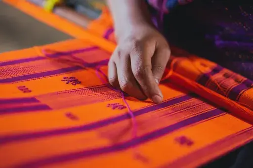 The brown-skinned hand of a textile worker is in focus, resting over loose threads on a bright orange and purple woven cloth.