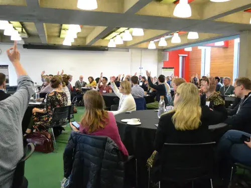 Groups of people sitting around tables listening to a speaker with their hands up