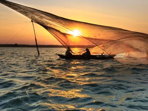 Two people sit silhouetted in a shallow boat on the water at sunset casting an enormous fishing net which floats like a diaphanous membrane above their heads © Pexels