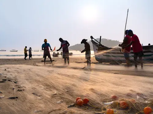 A dawn scene of fishermen working collaboratively, gathering nets on a sunlit coastal shore, with wooden boats docked nearby