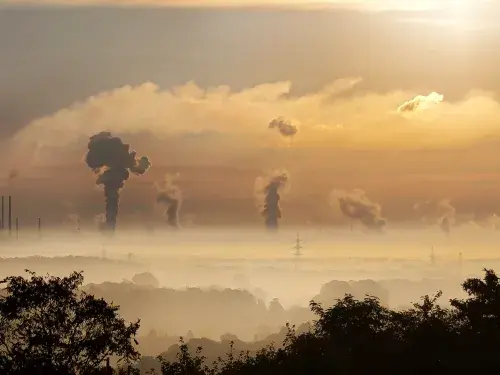 The sun rises over plumes of emissions gas from powerplants dotted along the horizon, which emanate into the sky beyond a smog which covers the trees and houses in the foreground