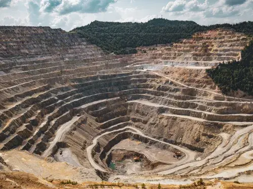 A deep, stepped mine site of brown and yellow rock and dust. Roads wind up the sides with mining vehicles moving up and down. Two yellow excavators sit at the bottom next to a pool of blue water. Dense forest mantles the upper edge and right hand shoulder of the site, all under a bright sunny sky with white clouds.