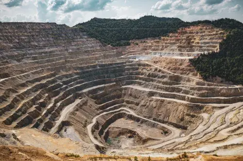 A deep, stepped mine site of brown and yellow rock and dust. Roads wind up the sides with mining vehicles moving up and down. Two yellow excavators sit at the bottom next to a pool of blue water. Dense forest mantles the upper edge and right hand shoulder of the site, all under a bright sunny sky with white clouds.