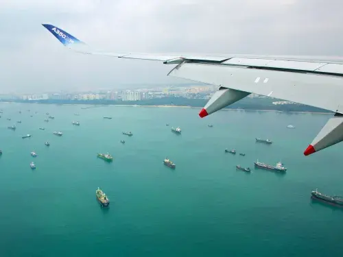View from an aircraft wing of a number of container ships anchored off a coastline featuring both forest and high rise buildings in a brightly coloured ocean © RSB