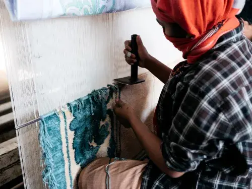 Person with an orange head scarf works on a white and blue rug © GoodWeave International
