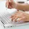 Person with their sleeves rolled up typing on a computer, holding a silver pen in one hand © Pexels