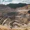 A deep, stepped mine site of brown and yellow rock and dust. Roads wind up the sides with mining vehicles moving up and down. Two yellow excavators sit at the bottom next to a pool of blue water. Dense forest mantles the upper edge and right hand shoulder of the site, all under a bright sunny sky with white clouds.