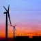 The silhouettes of four wind turbines stand against a blue sky at dusk with an orange sunset on the horizon